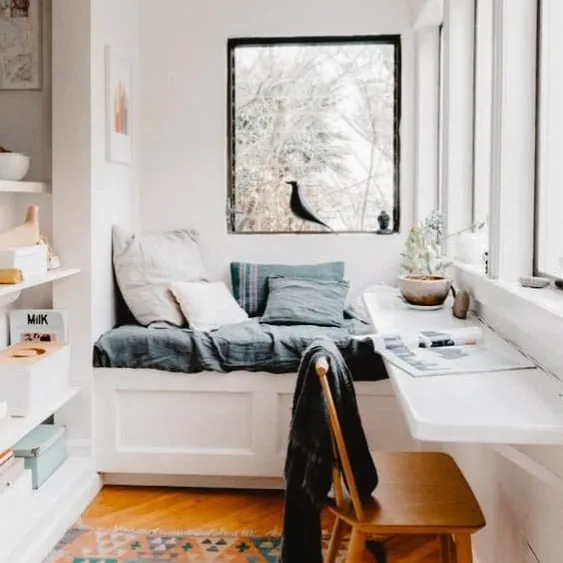 Cozy and minimalist reading nook with a daybed and built-in desk, surrounded by bookshelves and natural light from large windows, creating a peaceful retreat.