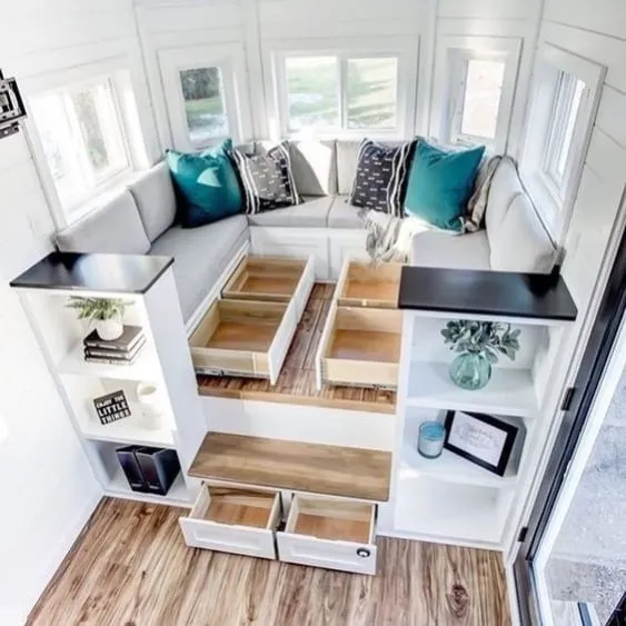 Bright and airy tiny house living area with built-in underfloor storage drawers, a cozy L-shaped couch, and modern shelving units.