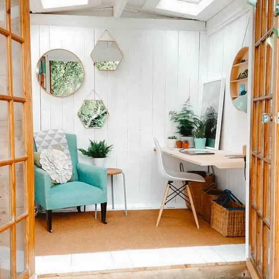 Cozy She Shed with a teal armchair, a small desk, and decorative mirrors on white walls, featuring natural light from skylights.