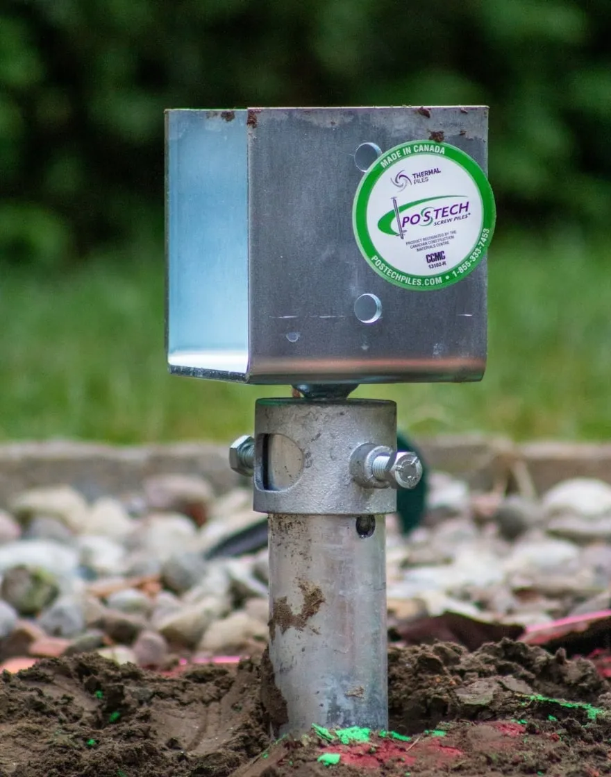 A close-up of a Postech screw pile, featuring a galvanized steel post installed securely into the ground with a metal bracket attached at the top. The Postech logo and branding are visible on the bracket.