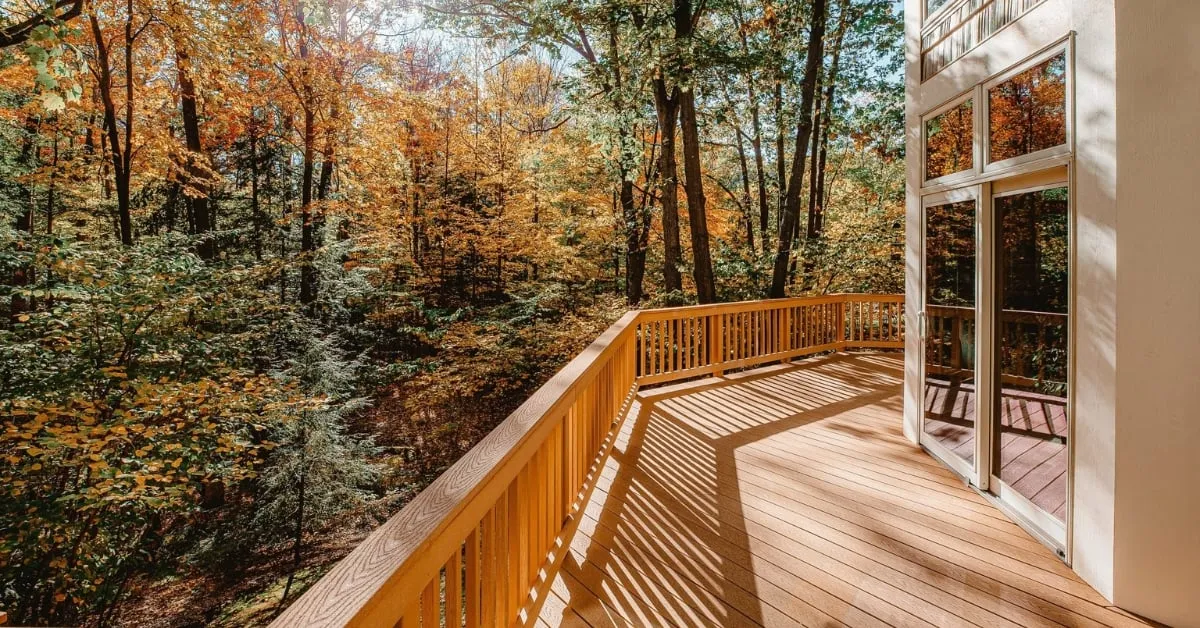 Patio en bois surplombant une forêt aux couleurs d'automne, avec de grandes fenêtres vitrées donnant sur l'intérieur.